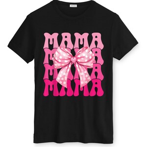 May include: Black t-shirt featuring the word "MAMA" in pink, repeated three times. A pink bow with white polka dots is centered on the design. The shirt has a crew neck and short sleeves.