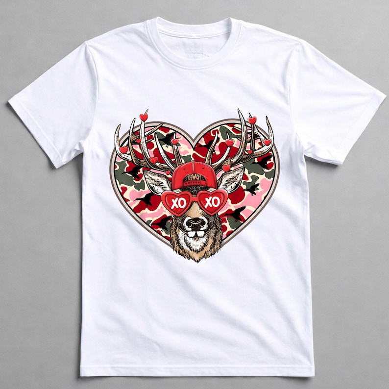 May include: White t-shirt featuring a deer graphic with a red cap and heart-shaped sunglasses that read "XO." The deer is set against a heart-shaped camouflage background, with small red hearts on the antlers. A fun and festive design.