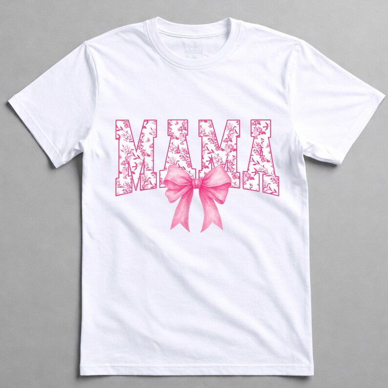 May include: White t-shirt featuring the word "MAMA" in pink floral print lettering and a pink bow. The t-shirt has a crew neck and is likely made of cotton or a similar material. The design is centrally placed on the front of the garment.