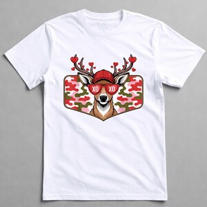 May include: White t-shirt featuring a deer graphic. The deer wears a red cap and heart-shaped sunglasses with "XO" on the lenses. Its antlers are adorned with red hearts. The graphic is set against a camouflage background.
