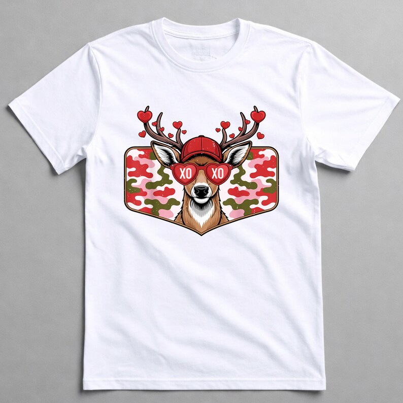 May include: White t-shirt featuring a deer graphic. The deer wears a red cap and heart-shaped sunglasses with "XO" on the lenses. Its antlers are adorned with red hearts. The graphic is set against a camouflage background.