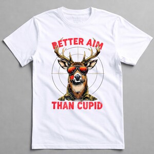 May include: White t-shirt featuring a deer wearing sunglasses and a military-style jacket, with the text "BETTER AIM THAN CUPID" in red. The deer is centered within a target reticle.