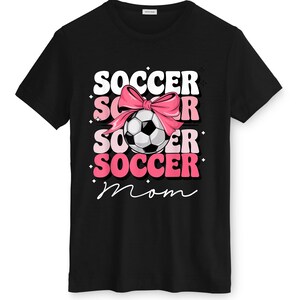May include: Black t-shirt with a graphic design featuring the words "SOCCER MOM" in white and pink, a soccer ball, and a pink bow. The soccer ball is black and white.