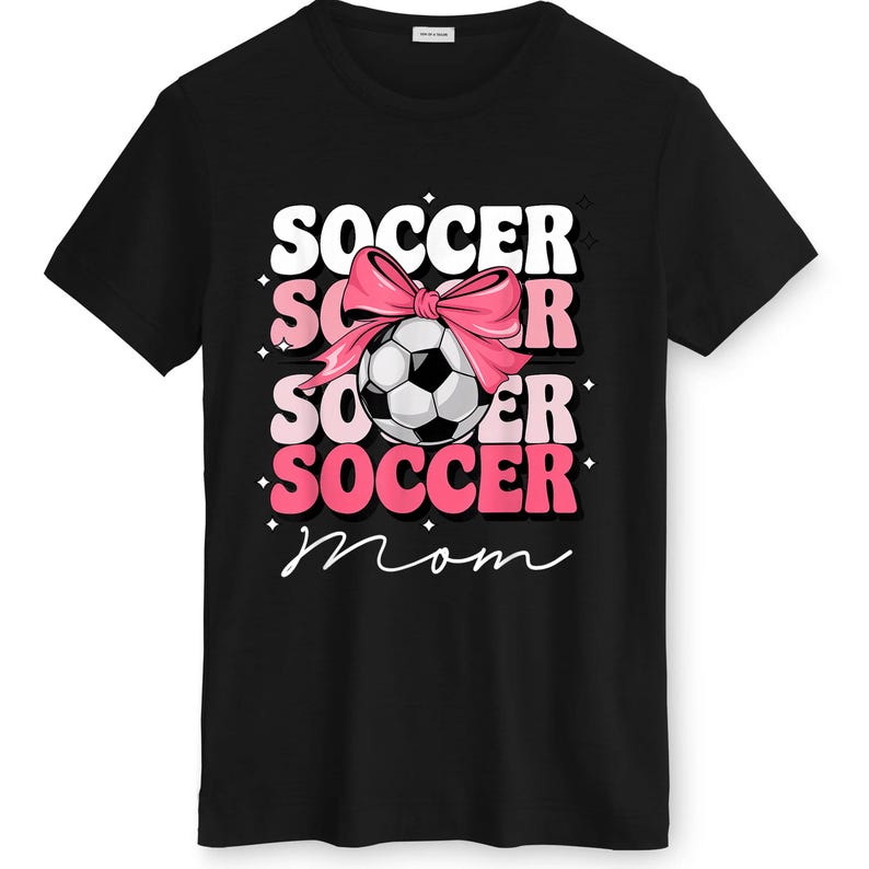 May include: Black t-shirt with a graphic design featuring the words "SOCCER MOM" in white and pink, a soccer ball, and a pink bow. The soccer ball is black and white.