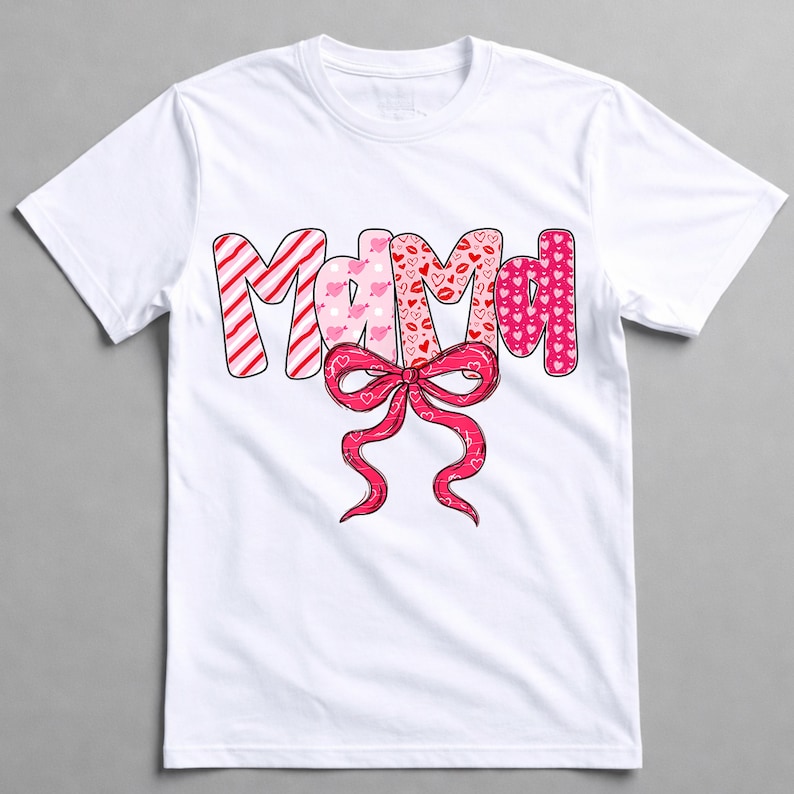 May include: White t-shirt featuring the word "Mama" in a colourful, patterned design. The letters are in pink and red with patterns of stripes, hearts, and lips. A pink bow is below the word.