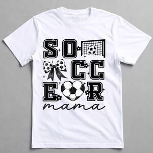 May include: White t-shirt with a black and white soccer-themed graphic. The design includes the word "SOCCER" with a soccer ball bow, a soccer net, and a heart-shaped soccer ball. The word "mama" is written in cursive.