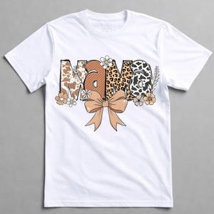 May include: White t-shirt with the word "MAMA" in a decorative design. The letters feature cow print, leopard print, and floral patterns. A tan bow and floral accents are below the word.