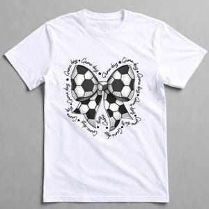 May include: White t-shirt featuring a soccer ball bow design. The bow is black and white, with the words "Game Day" and stars encircling the bow. A casual tee for soccer fans.