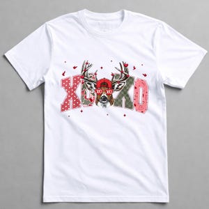 May include: White t-shirt featuring a deer wearing a red cap and antlers adorned with hearts. The shirt has the text "XO XO" in pink and green patterned letters, with small red hearts and birds scattered around the design.