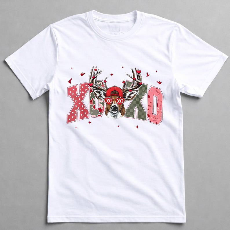 May include: White t-shirt featuring a deer wearing a red cap and antlers adorned with hearts. The shirt has the text "XO XO" in pink and green patterned letters, with small red hearts and birds scattered around the design.