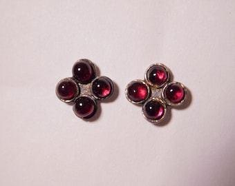 May include: A pair of stud earrings, each featuring a cluster of five deep red, round gemstones set in a silver-toned metal. The earrings are arranged in a four-petal flower shape.