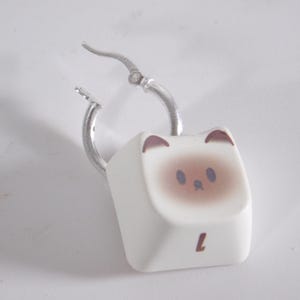May include: A single silver hoop earring with a white cat-shaped charm. The charm is a keyboard key with a cat face design and the letter "L" printed on it. The cat's face has brown accents.