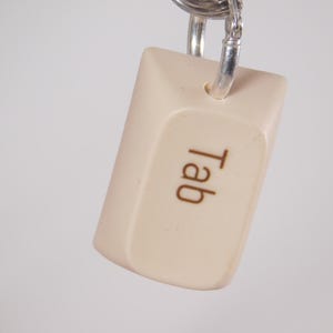 May include: A beige keycap-shaped keychain with the word "Tab" printed in brown. The keychain is attached to a silver keyring. The background is a soft, neutral color.