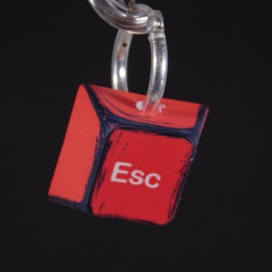 May include: A red cube-shaped keychain with the word "Esc" in white. The edges are outlined in dark blue with a brushstroke effect. The keychain is attached to a silver ring.