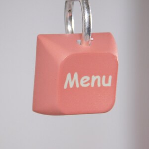 Könnte beinhalten: Ein rosa, würfelförmiger Anhänger mit dem Wort "Menu" in weißer Schrift. Der Anhänger wird von einem silbernen Metallring gehalten. Der Hintergrund ist eine weiche, neutrale Farbe.