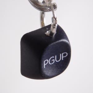 May include: A black keycap with the letters "PGUP" in white, attached to a silver keyring. The keycap is a cube shape with rounded edges. The background is plain white.