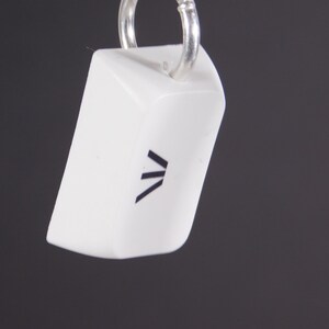 May include: A white, rectangular pendant with a silver ring. The pendant has a black graphic design on the front. The background is a solid dark gray.