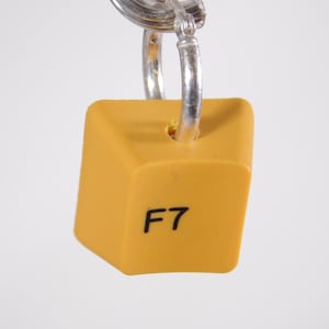 May include: Mustard yellow keycap keychain with the letter "F" and the number "7" printed in black. The keycap is attached to a silver-colored metal keyring. The background is a plain white.