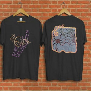 Puede incluir: Dos camisetas negras colgando de perchas de madera. Una camiseta tiene el texto "the Cure" en un diseño estilizado con acentos morados y dorados. La otra camiseta presenta un gráfico de pulpo con el texto "the Cure".