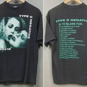 May include: Dark gray t-shirt featuring a black and white image of two women kissing, with the text "Bloody Kisses" and "Type O Negative." The back of the shirt has a list of items with the text "Type O Negative is to blame for:"