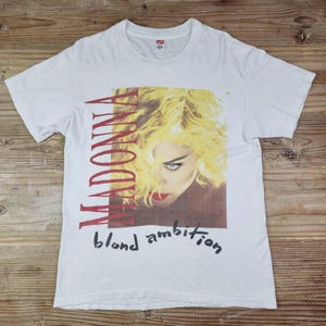 Puede incluir: Camiseta blanca con un retrato de Madonna con pelo rubio y pintalabios rojo. La palabra "MADONNA" está impresa en rojo, y "blond ambition" está escrita en guion negro debajo de la imagen. La camiseta es de material suave.