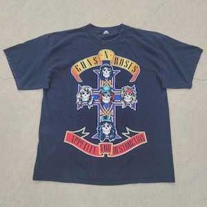 Puede incluir: Camiseta azul marino con el logo de Guns N' Roses y la frase "Appetite for Destruction". El diseño presenta una cruz con ilustraciones de calaveras. El nombre de la banda está en una pancarta naranja.