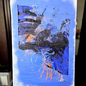 Könnte beinhalten: Abstrakte Malerei auf Leinwand mit blauem Hintergrund und schwarzem und orangefarbenem Design. Das Kunstwerk zeigt Spritzer, Linien und kreisförmige Formen. Die Leinwand ist auf einer Holzstaffelei montiert.