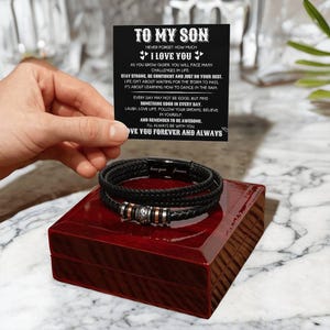 Peut inclure: Un bracelet en cuir tressé noir avec des perles argentées et cuivrées, posé sur un coffret en bois rouge. Une carte avec le texte "TO MY SON" et un message est tenue au-dessus du bracelet.