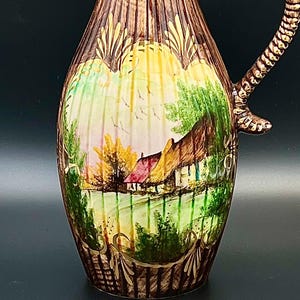 May include: A tall ceramic pitcher with a brown wood-grain texture and a ribbed surface. The front features a hand-painted scene of a cottage with a thatched roof, green trees, and a yellow sky. It has a curved handle with a textured, rope-like design.