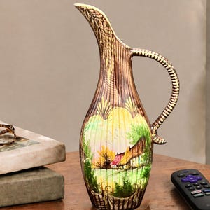 May include: A tall, slender ceramic pitcher featuring a textured, wood-grain finish on the neck and handle, with a hand-painted landscape scene on the body depicting a small cottage nestled among green trees and yellow foliage.