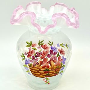 May include: A clear glass vase with a pink-edged, ruffled rim. The vase is decorated with a hand-painted design of a basket overflowing with red and purple flowers and green leaves. The vase has a rounded shape.