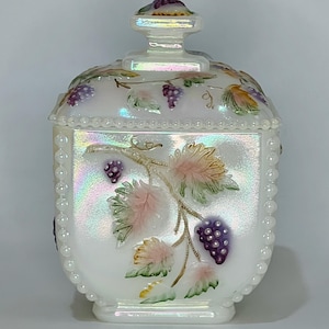 May include: White, iridescent glass jar with a lid, featuring a grape vine design. The jar has a square shape with rounded edges, a beaded border, and a decorative knob on the lid. The grapes are purple, and the leaves are green and pink.