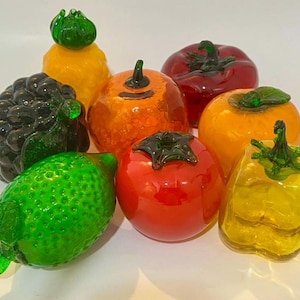 May include: A collection of eight glass fruit and vegetable figurines including a textured green lime, a dark purple grape cluster, a yellow pineapple, a bright orange citrus, a red tomato, a yellow bell pepper, and a deep red bell pepper.