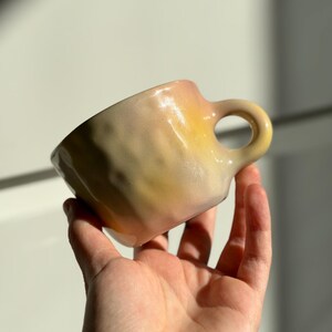May include: A handmade ceramic mug with a unique, textured surface. The mug features a gradient of colors, transitioning from a soft pink at the rim to a sunny yellow at the base. It has a rounded handle and a slightly irregular shape, suggesting it's a piece of artisan pottery.