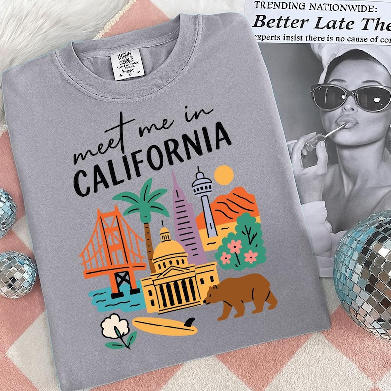 K&ouml;nnte beinhalten: Ein graues T-Shirt mit dem Schriftzug "meet me in CALIFORNIA" und farbenfrohen Illustrationen kalifornischer Wahrzeichen, darunter die Golden Gate Bridge, eine Palme und ein B&auml;r. Das Shirt hat auch ein kleines Etikett oben links.