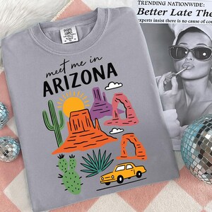 K&ouml;nnte beinhalten: Graues T-Shirt mit dem Text "meet me in ARIZONA" und farbenfrohen Illustrationen einer W&uuml;stenlandschaft, einschlie&szlig;lich Sonne, Kakteen und Felsformationen. Ein gelbes Auto ist ebenfalls abgebildet.