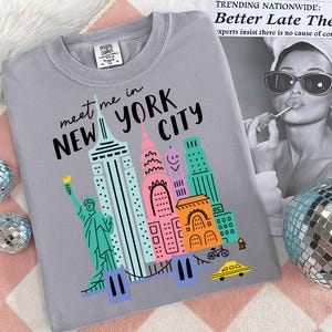 K&ouml;nnte beinhalten: Ein graues T-Shirt mit einer bunten Grafik von New Yorker Wahrzeichen, darunter die Freiheitsstatue und Wolkenkratzer. Der Text "meet me in NEW YORK CITY" ist &uuml;ber der Illustration aufgedruckt. Das Shirt liegt auf einer rosa-wei&szlig;en Oberfl&auml;che.
