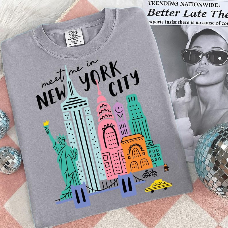 K&ouml;nnte beinhalten: Ein graues T-Shirt mit einer bunten Grafik von New Yorker Wahrzeichen, darunter die Freiheitsstatue und Wolkenkratzer. Der Text "meet me in NEW YORK CITY" ist &uuml;ber der Illustration aufgedruckt. Das Shirt liegt auf einer rosa-wei&szlig;en Oberfl&auml;che.