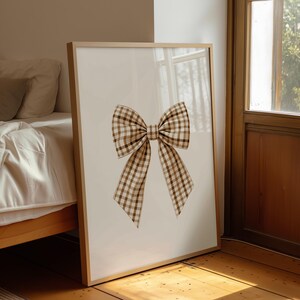 Può includere: Stampa incorniciata con un'illustrazione di un grande fiocco. Il fiocco è realizzato con un motivo a quadri vichy marrone e crema. L'opera d'arte è esposta in una cornice di legno chiaro, appoggiata a un muro vicino a una finestra e un letto.