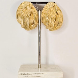 May include: A pair of gold-toned earrings featuring a textured, woven metal design shaped into a rounded knot. The earrings have a polished, reflective finish and are displayed on a vertical metal stand with a rectangular base.