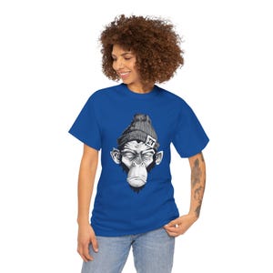Könnte beinhalten: Ein königsblaues T-Shirt mit einer Schwarz-Weiß-Illustration eines Affengesichts, das eine graue Mütze mit den Buchstaben "HT" trägt. Der Affe hat geschlossene Augen und einen neutralen Ausdruck. Das Shirt ist ein Rundhals-Stil.