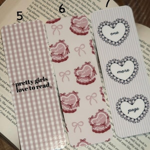May include: Three decorative bookmarks with different designs. One bookmark has a pink gingham pattern with the text "pretty girls love to read." Another features heart-shaped designs with bows. The third has heart shapes with the words "one," "more," and "page."