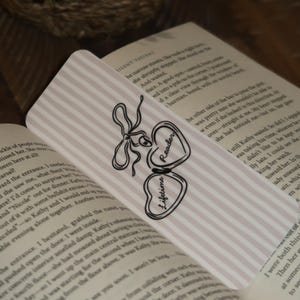 May include: A decorative bookmark with a pink and white striped background. The bookmark features a black and white bow design and two heart-shaped charms. One charm says "Reader" and the other says "Lifetime".