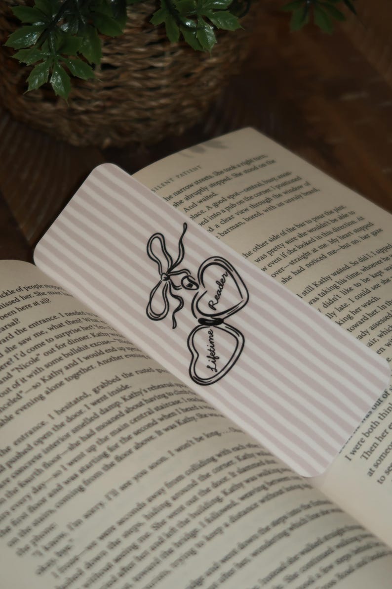 May include: A decorative bookmark with a pink and white striped background. The bookmark features a black and white bow design and two heart-shaped charms. One charm says "Reader" and the other says "Lifetime".