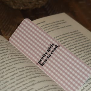 May include: A pink and white gingham bookmark with the text "pretty girls love to read" in black script. The bookmark is placed in an open book with text visible on the pages.