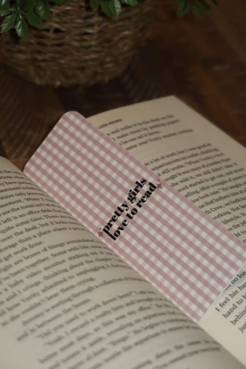 May include: A pink and white gingham bookmark with the text "pretty girls love to read" in black script. The bookmark is placed in an open book with text visible on the pages.