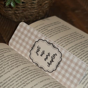 May include: A beige and white gingham bookmark with the text "just one more chapter" in a decorative frame, placed in an open book. The bookmark adds a touch of style to the reading experience.