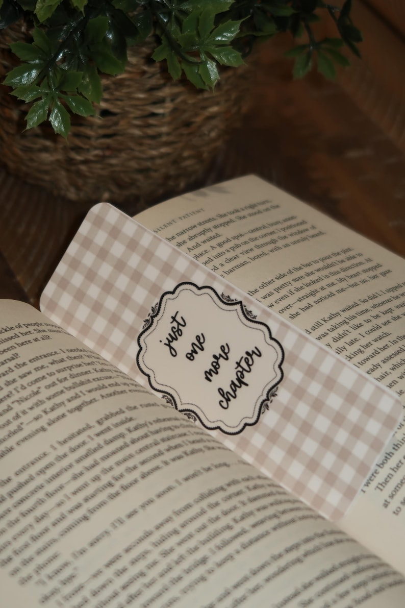 May include: A beige and white gingham bookmark with the text "just one more chapter" in a decorative frame, placed in an open book. The bookmark adds a touch of style to the reading experience.