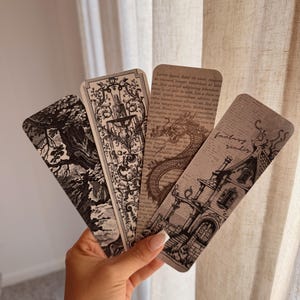 May include: Four vintage-style bookmarks with intricate black and white illustrations. Designs include a tree, ornate fountain, dragon, and a building with the text "fantasy reader". The bookmarks are held in a hand.