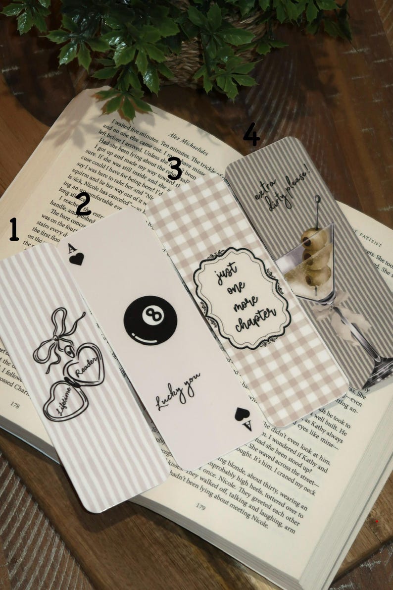 May include: A collection of decorative bookmarks with various designs, including hearts, a pool ball, and the text "Lucky you." One bookmark says "just one more chapter." The bookmarks are on top of an open book.